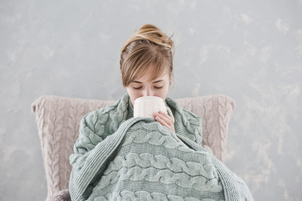 Woman with blond hair sitting with knees bent up in front of her, holding a white cup of hot drink wrapped with light green blanket at home. Gray background.